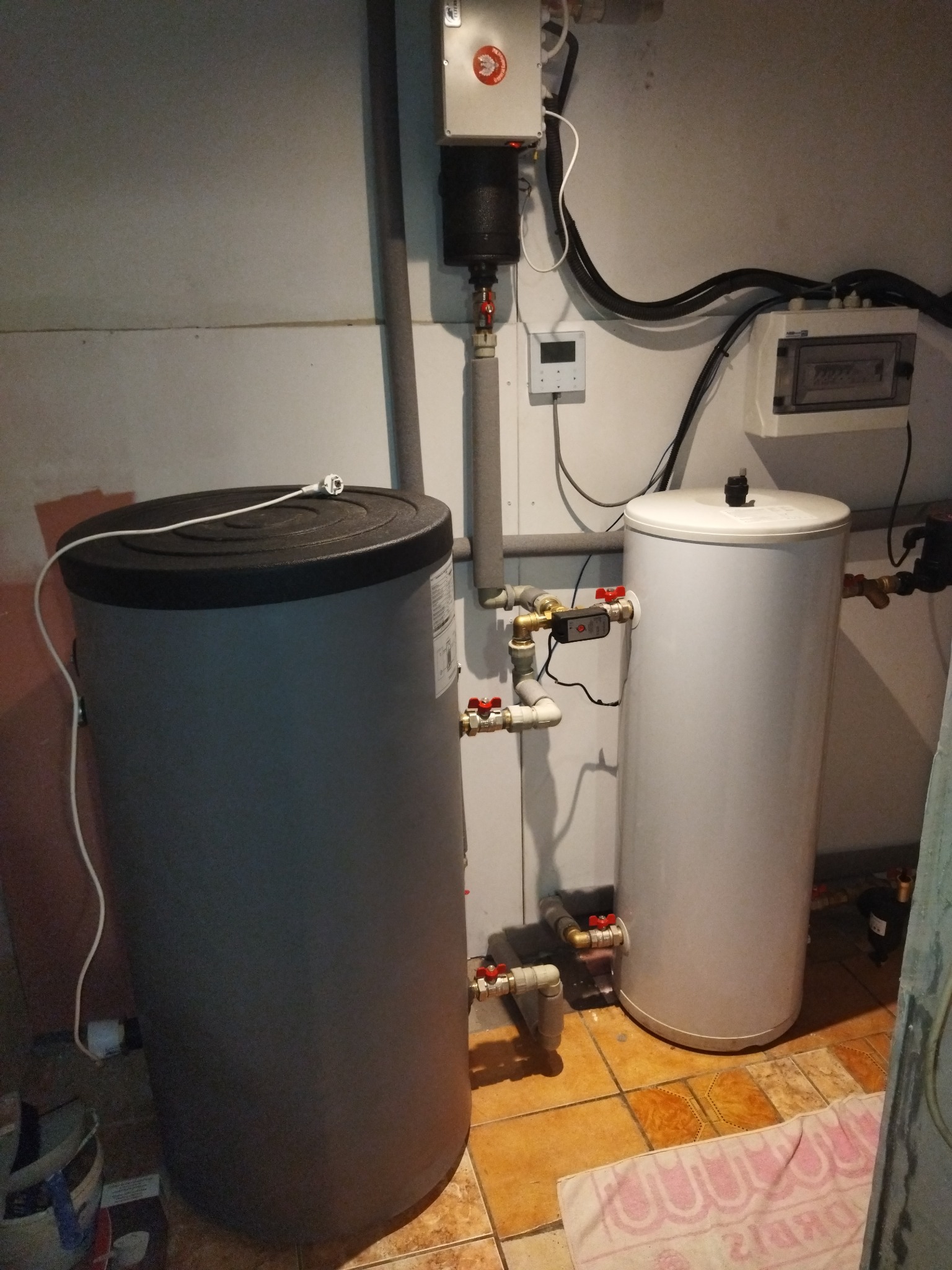 Dwa zbiorniki akumulacyjne ciepła w kotłowni, rury z zaworami, sterownik i zabezpieczenia elektryczne na ścianie.