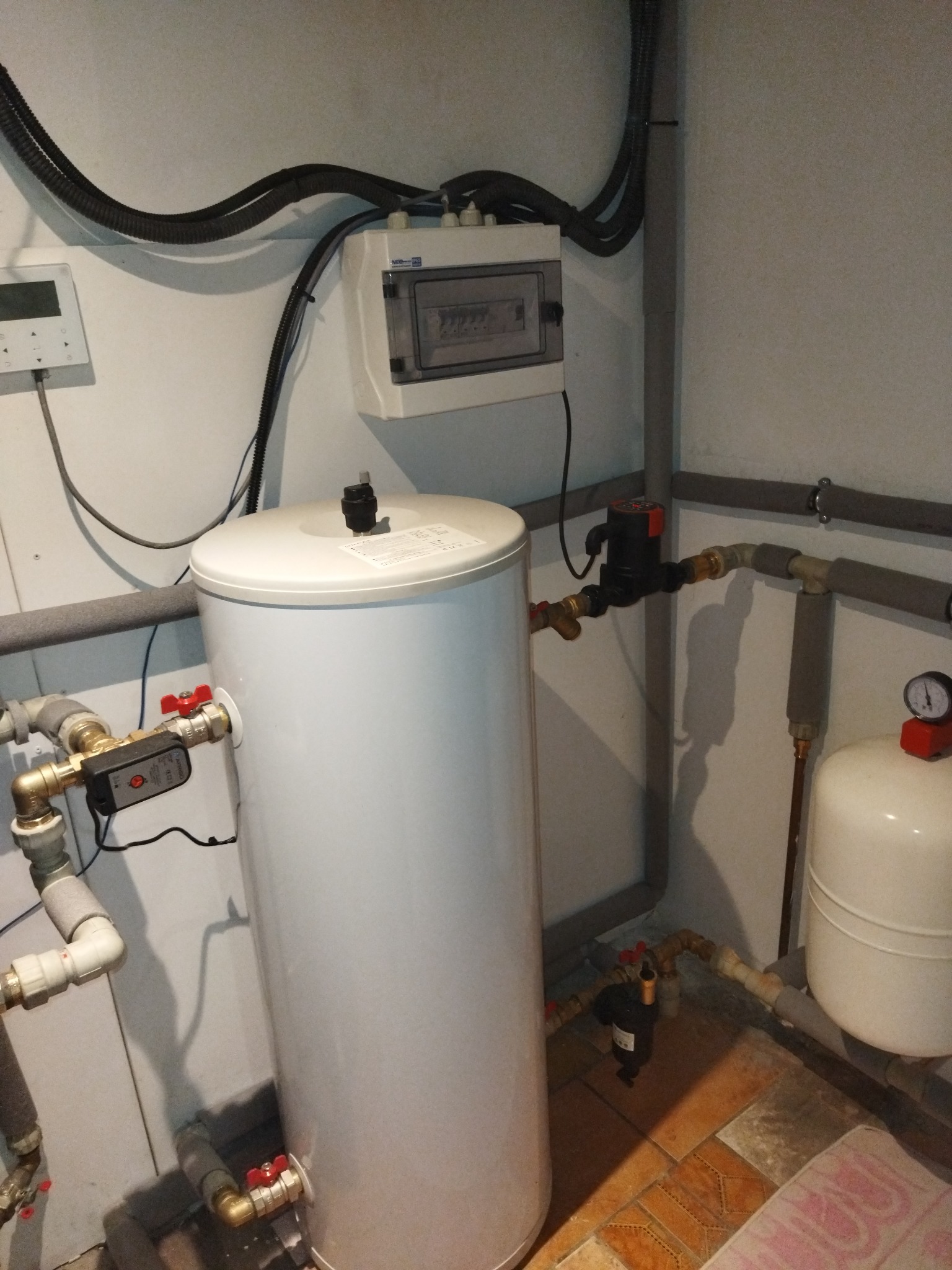 Biały zasobnik ciepłej wody użytkowej z podłączonymi rurami i zaworami w kotłowni, widoczna instalacja elektryczna i zabezpieczenia.