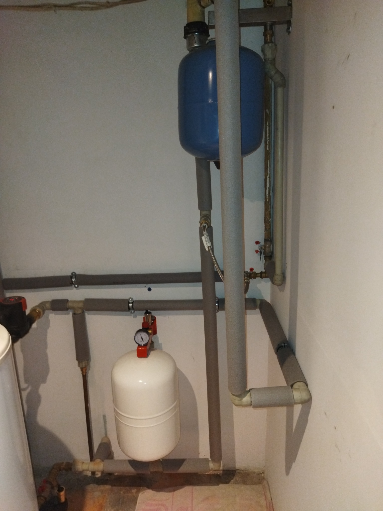 Instalacja grzewcza w piwnicy: niebieski zbiornik wyrównawczy nad białym, rury z izolacją termiczną, manometr na górze zbiornika.