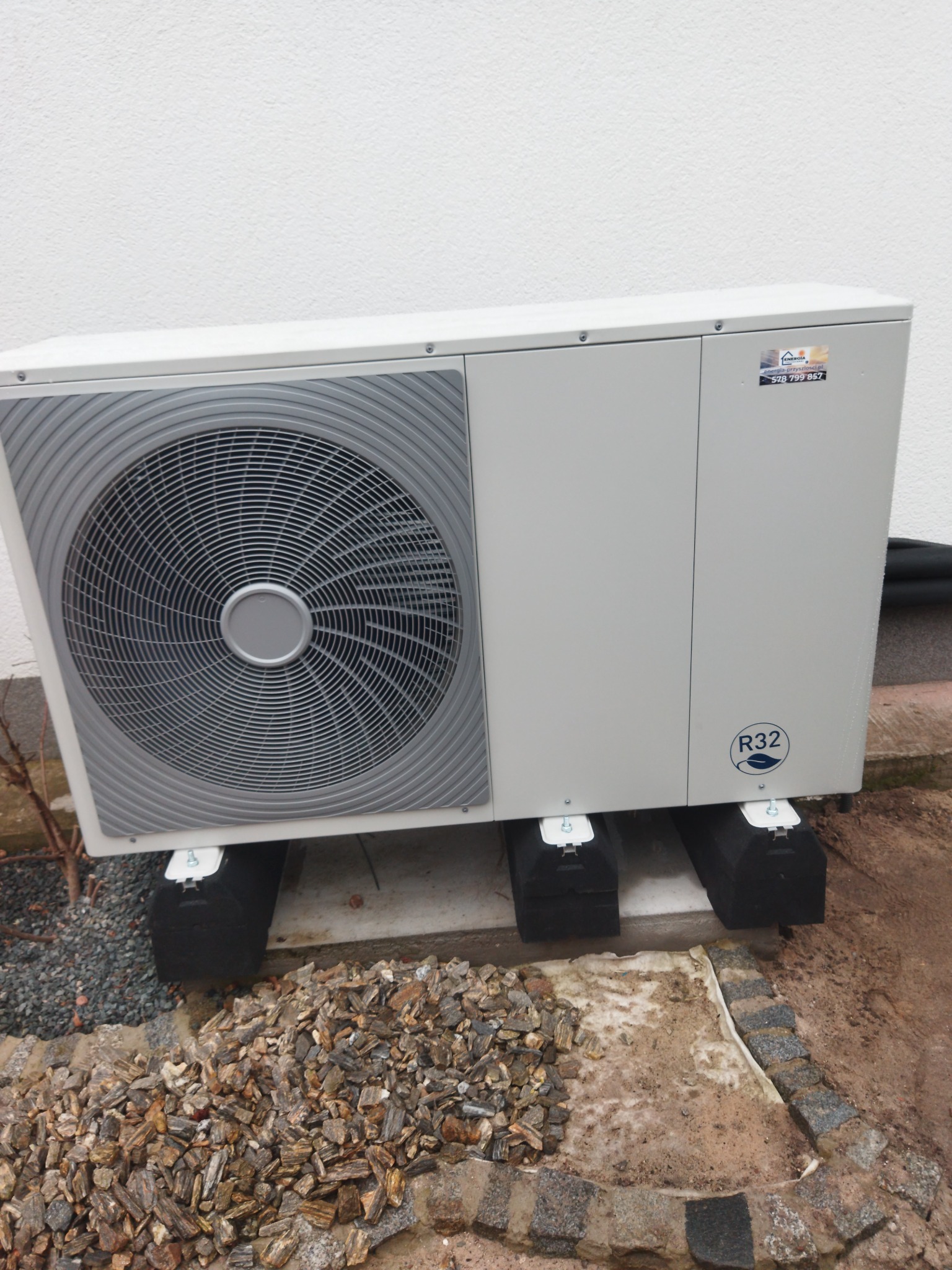 Zewnętrzna jednostka grzewcza na czarnych podkładkach, widoczny wentylator i naklejka informacyjna R32, otoczenie wyłożone kamieniami.