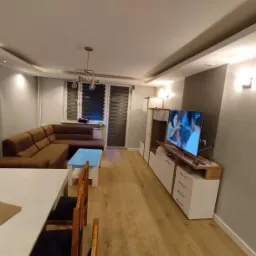 Jasne wnętrze pokoju dziennego z narożną sofą, białym stolikiem kawowym z błękitnym blatem i meblościanką z telewizorem, oświetlone listwami LED na suficie.