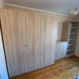 Jasna, drewnopodobna szafa wnękowa z czterema drzwiami i dodatkowymi szufladami oraz regałem, na tle drewnianej podłogi w trakcie montażu.