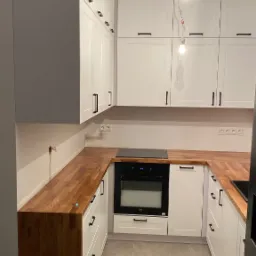 Biała kuchnia z drewnianym blatem, czarnymi uchwytami i niedokończonym oświetleniem sufitowym.