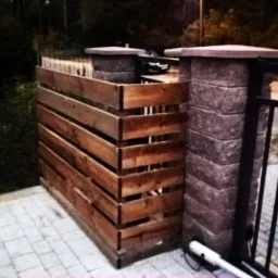 Narożny fragment drewnianego ogrodzenia z poziomych desek, przylegający do murowanego słupka z betonową czapą, widoczny fragment metalowej bramy.