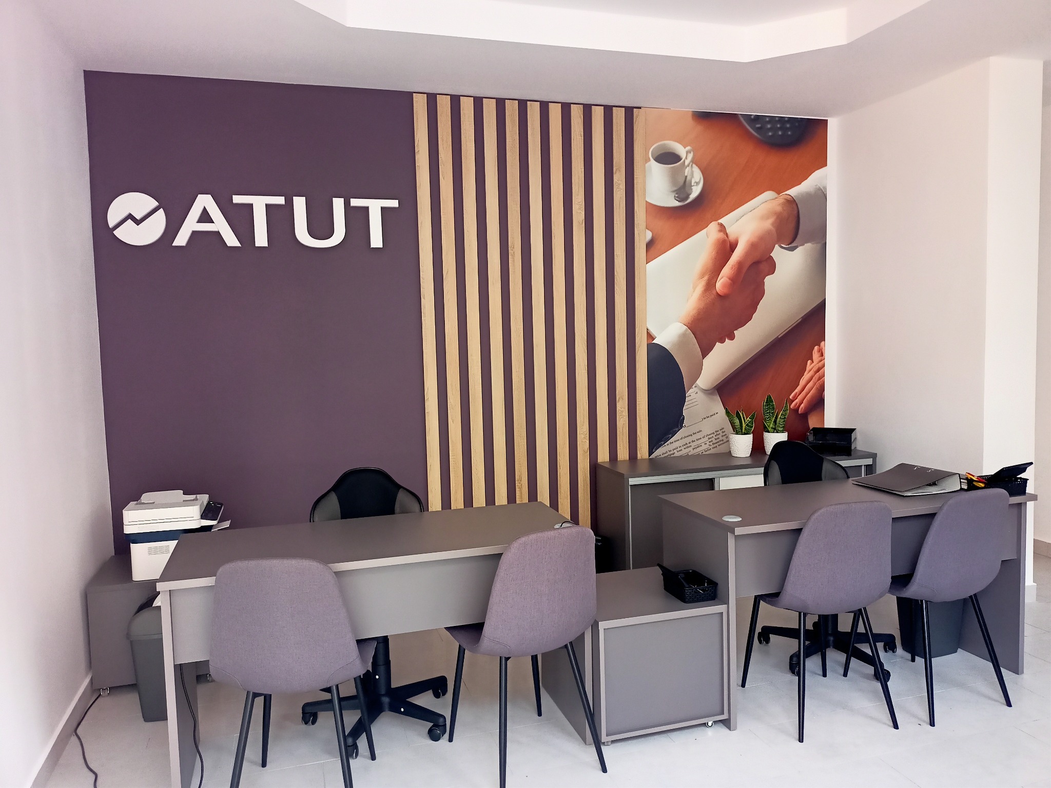 Biuro z logo 'ATUT' na ścianie, drewnianymi lamelami i fototapetą z uściskiem dłoni nad filiżanką kawy. Dwa biurka z szarymi krzesłami i wyposażeniem biurowym.