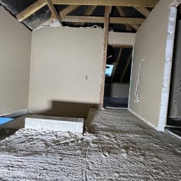 Anna&Piotr Tynk - Poddasze w trakcie remontu, ściany wstępnie przygotowane, widoczne przewody elektryczne, podłoga pokryta pyłem, drewniana konstrukcja dachu. W tle widoczne okno dachowe.