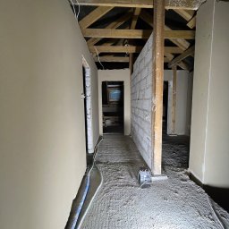 Anna&Piotr Tynk - Wnętrze poddasza w trakcie prac wykończeniowych; ściany pokryte świeżą warstwą tynku, widoczne drewniane elementy konstrukcyjne dachu, podłoga pokryta warstwą pyłu i resztek materiałów budowlanych,...