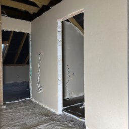 Anna&Piotr Tynk - Świeżo położone tynki maszynowe w surowym wnętrzu poddasza, widoczne drewniane belki stropowe, wystające przewody elektryczne i niedokończone ościeżnice.