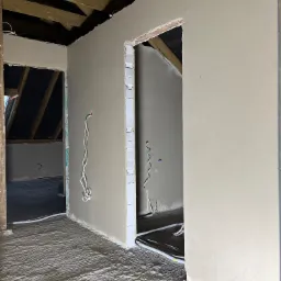 Świeżo położone tynki maszynowe w surowym wnętrzu poddasza, widoczne drewniane belki stropowe, wystające przewody elektryczne i niedokończone ościeżnice.