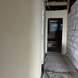 Anna&Piotr Tynk - Wnętrze poddasza w trakcie prac wykończeniowych: ściana z nałożoną warstwą wygładzającą, surowy mur z pustaków, drewniana konstrukcja dachu, okno dachowe w oddali, widoczne przewody elektryczne.