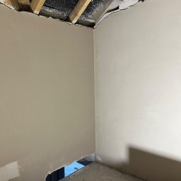 Anna&Piotr Tynk - Wnętrze w trakcie prac wykończeniowych; widoczne surowe drewniane belki stropowe z resztkami białego pyłu, częściowo wykończone ściany w jasnym odcieniu beżu, oraz fragment schodów z widocznym...