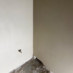 Anna&Piotr Tynk - Wnętrze po nałożeniu warstwy wykończeniowej na ścianę, widoczny fragment ściany z wystającym kablem elektrycznym oraz surowa podłoga w narożniku pomieszczenia.