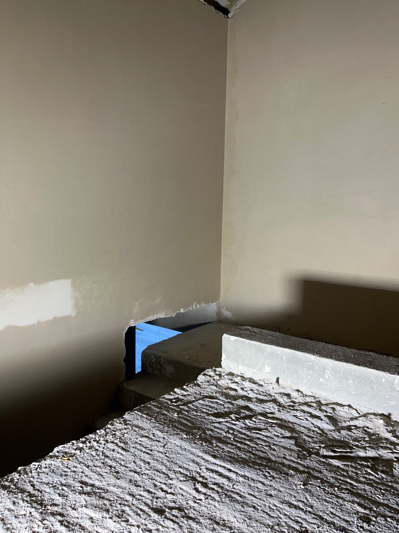 Wnętrze w trakcie prac wykończeniowych, widoczna ściana z fragmentem świeżo nałożonego materiału, surowy betonowy element konstrukcyjny w dolnej części kadru, niebieskie światło w tle.