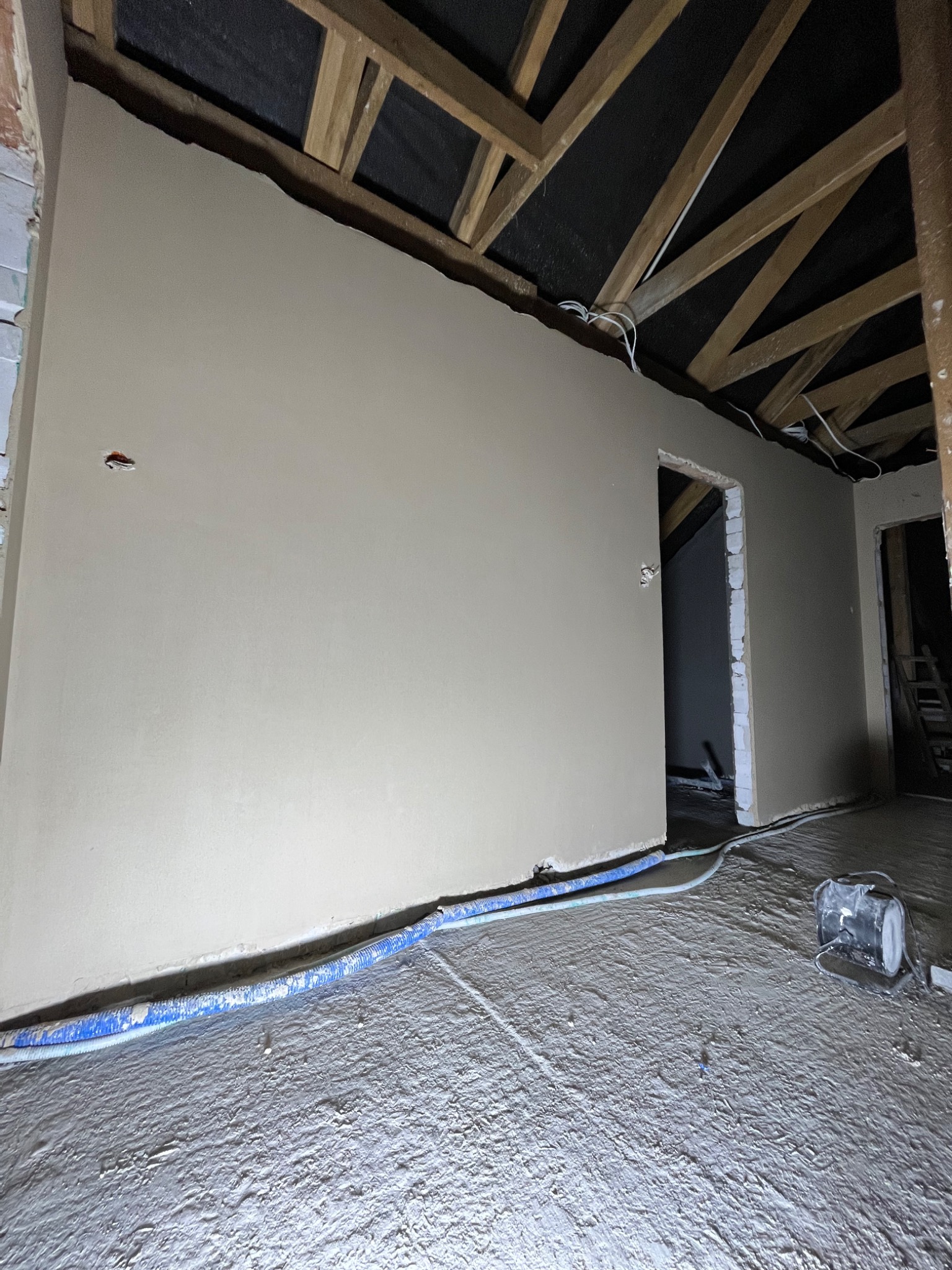 Wnętrze w trakcie remontu: ściana pokryta świeżo nałożoną warstwą wykończeniową, widoczne kable elektryczne wystające ze ściany, drewniana konstrukcja dachu oraz niebieskie rury przy podłodze.