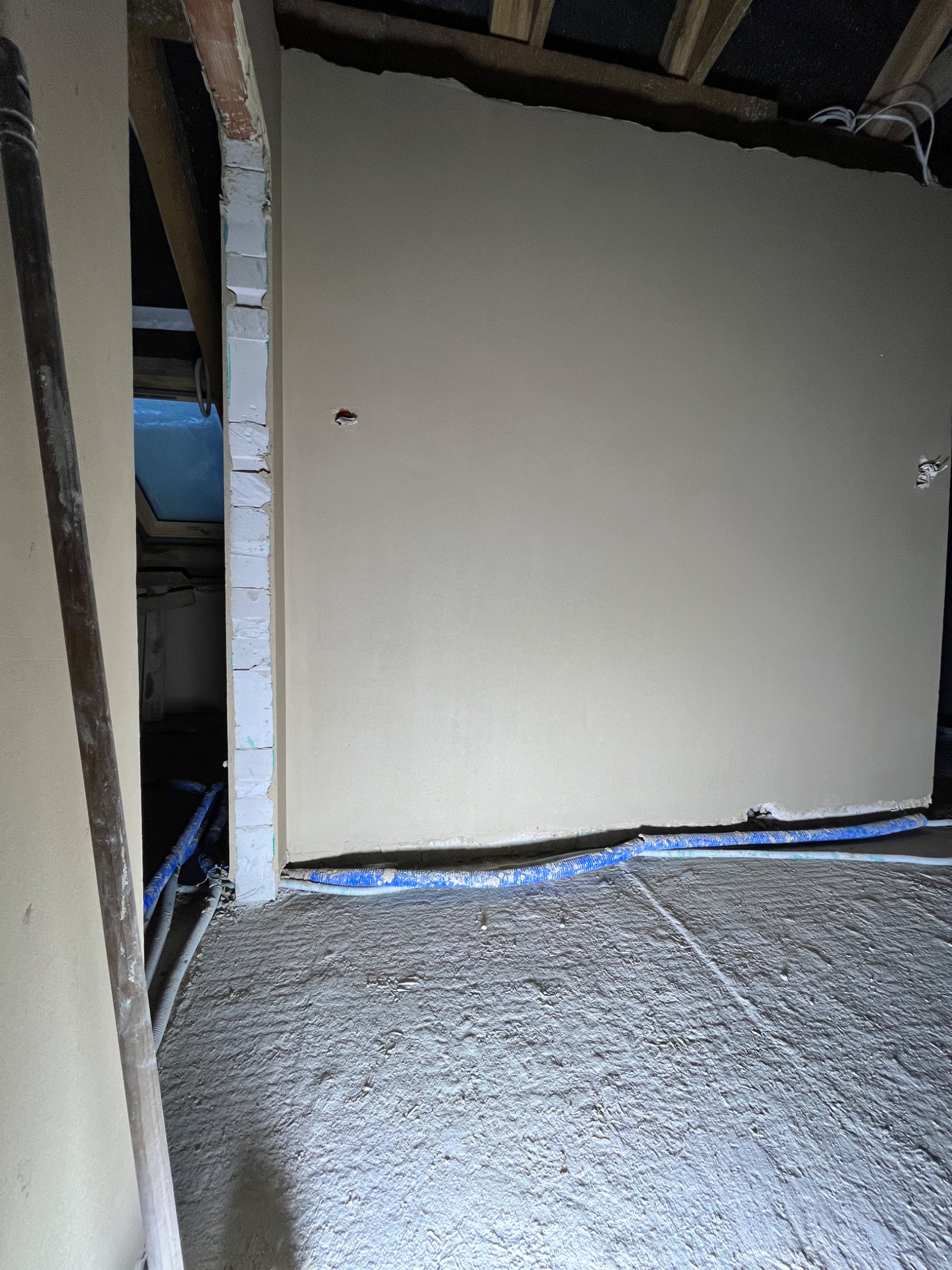 Świeżo nałożona warstwa wykończeniowa na ścianie, z widocznymi niebieskimi przewodami wzdłuż podłogi i drewnianą łatą opartą o ścianę po lewej stronie, ukazująca etap prac wykończeniowych...