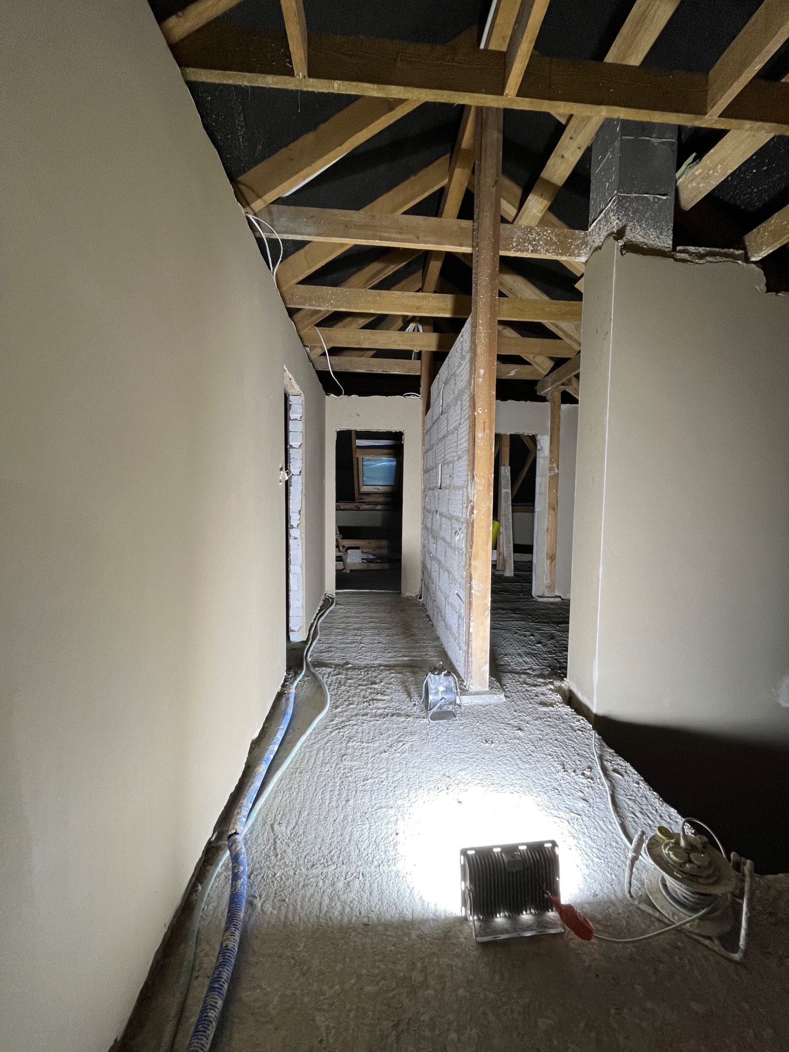 Wnętrze w trakcie prac wykończeniowych: ściany po nałożeniu gładzi, widoczne drewniane elementy konstrukcji dachu, prowizoryczne oświetlenie halogenowe na podłodze.