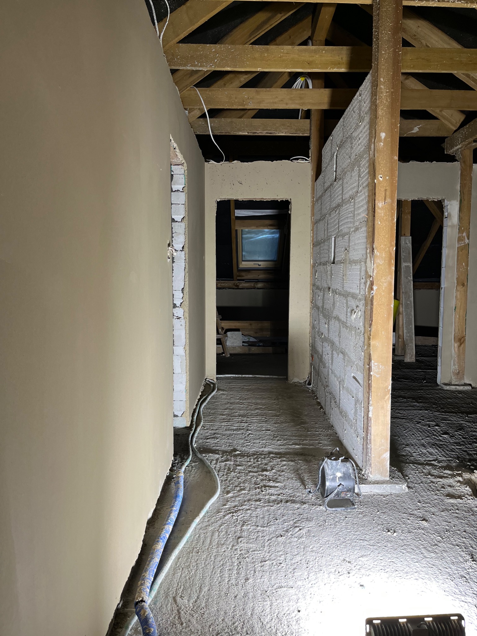 Wnętrze poddasza w trakcie prac wykończeniowych, ściana po lewej pokryta świeżo nałożoną, jasną warstwą, surowy beton na podłodze, widoczne fragmenty drewnianej konstrukcji dachu i lampa halogenowa...