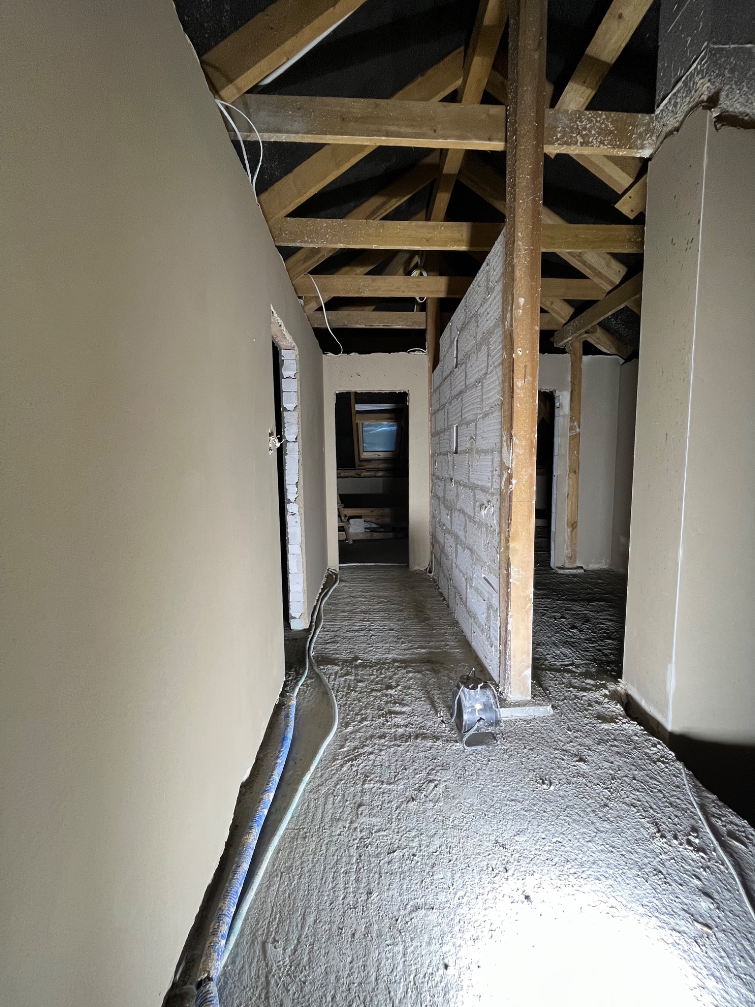 Wnętrze poddasza w trakcie prac wykończeniowych; ściany pokryte świeżą warstwą tynku, widoczne drewniane elementy konstrukcyjne dachu, podłoga pokryta warstwą pyłu i resztek materiałów budowlanych,...