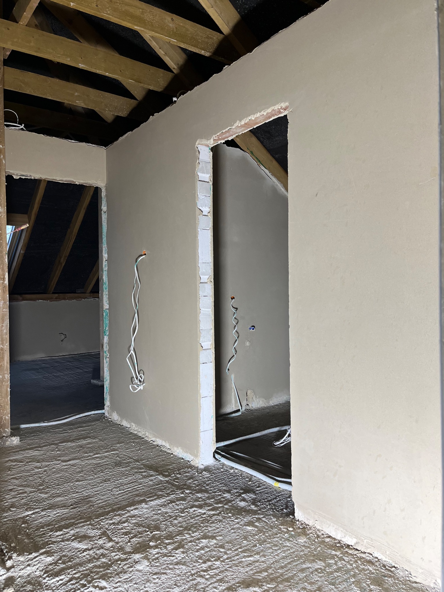 Świeżo położone tynki maszynowe w surowym wnętrzu poddasza, widoczne drewniane belki stropowe, wystające przewody elektryczne i niedokończone ościeżnice.