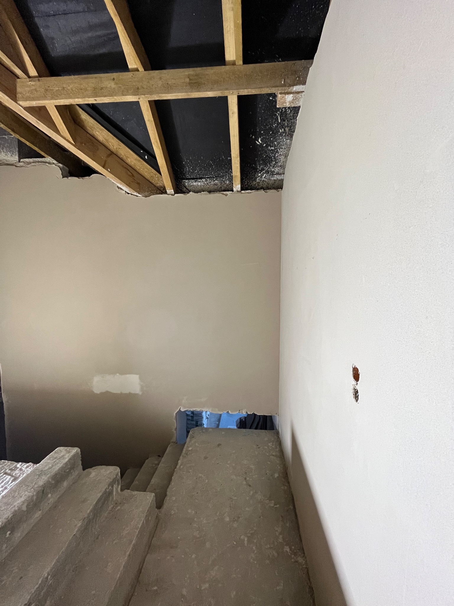 Surowe wnętrze w trakcie remontu: widoczne fragmenty drewnianej konstrukcji stropu, częściowo wykończone ściany z widocznymi otworami na instalacje elektryczne oraz betonowe schody prowadzące w dół.