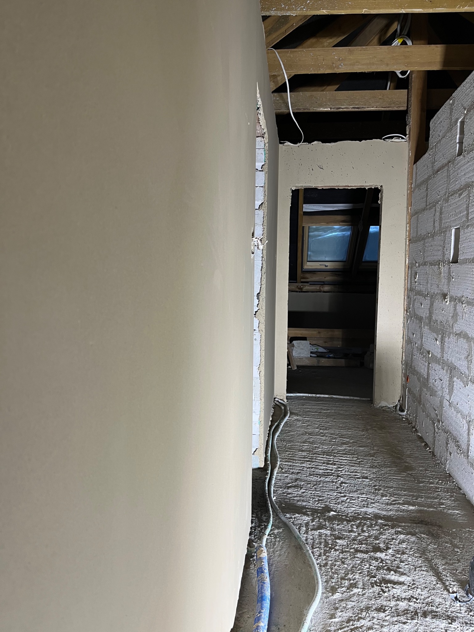 Wnętrze poddasza w trakcie prac wykończeniowych: ściana z nałożoną warstwą wygładzającą, surowy mur z pustaków, drewniana konstrukcja dachu, okno dachowe w oddali, widoczne przewody elektryczne.