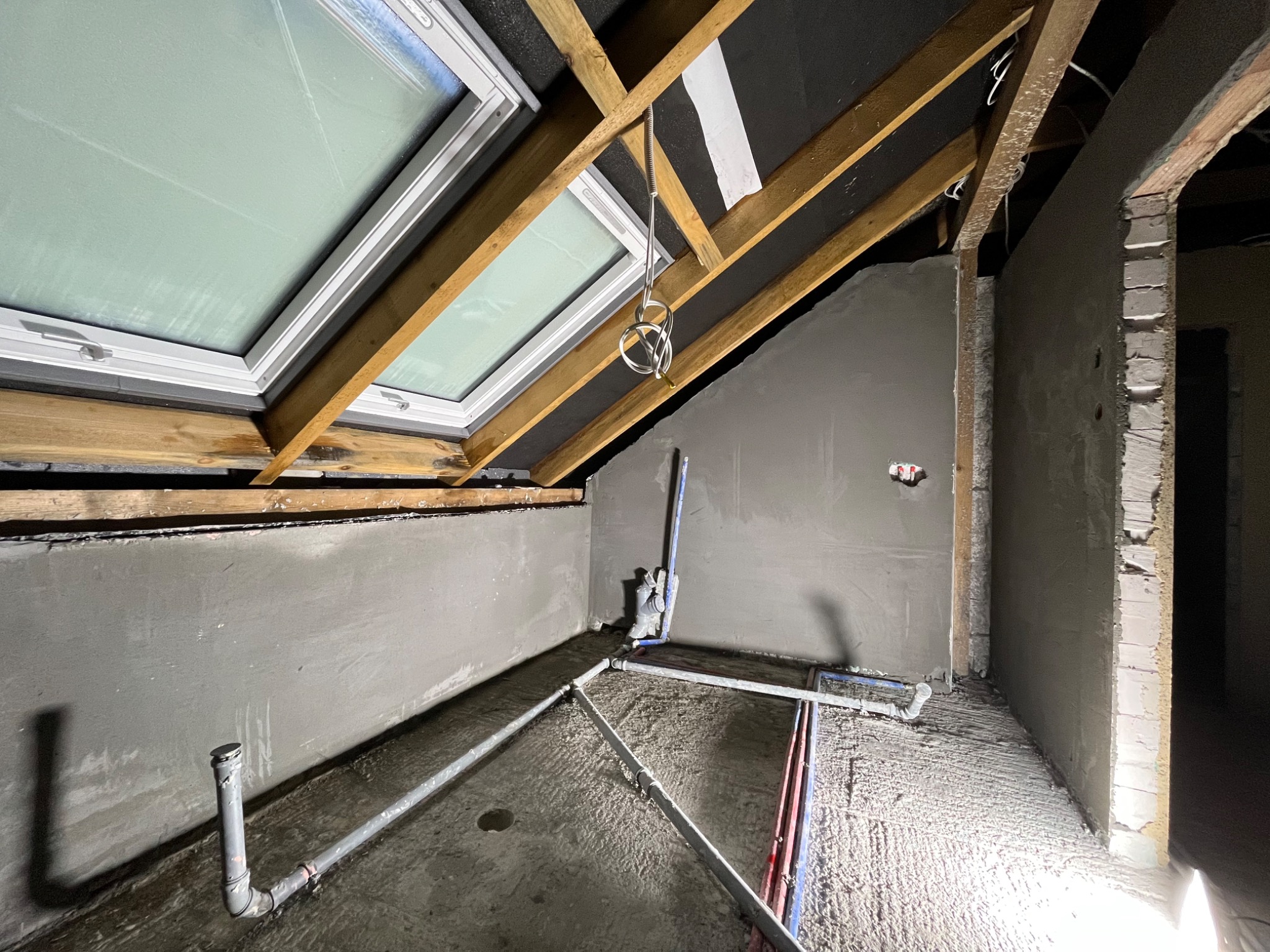 Poddasze w trakcie prac wykończeniowych: świeżo otynkowane ściany pod skosem z widocznym oknem dachowym, instalacja wodno-kanalizacyjna na surowym podłożu, drewniana konstrukcja dachu z czarną...