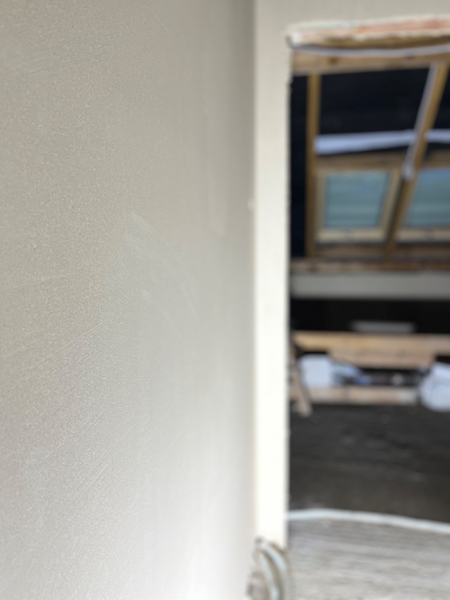 Świeżo nałożona, jasna gładź szpachlowa na ścianie, widoczne delikatne ślady po szpachli, w tle zamazane okno dachowe i elementy konstrukcyjne poddasza.