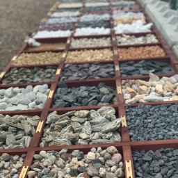 Monolit Invest - Ekspozycja różnorodnych kamieni dekoracyjnych i kruszyw w drewnianych przegródkach, z etykietami opisującymi rodzaje, prezentowana z perspektywy z góry, ukazująca bogactwo kolorów i faktur.