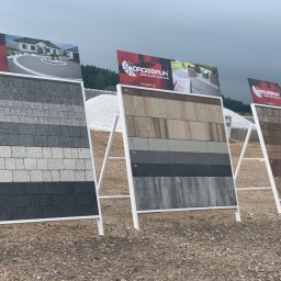 Monolit Invest - Ekspozycja trzech paneli z różnymi wzorami i kolorami kostki brukowej firmy Drogbruk na żwirowym podłożu, z widocznymi plakatami reklamowymi z logo i zdjęciami aranżacji.