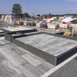 Ekspozycja różnych wzorów nawierzchni na placu budowlanym, z paletami materiałów w tle i wiatrakiem w oddali.