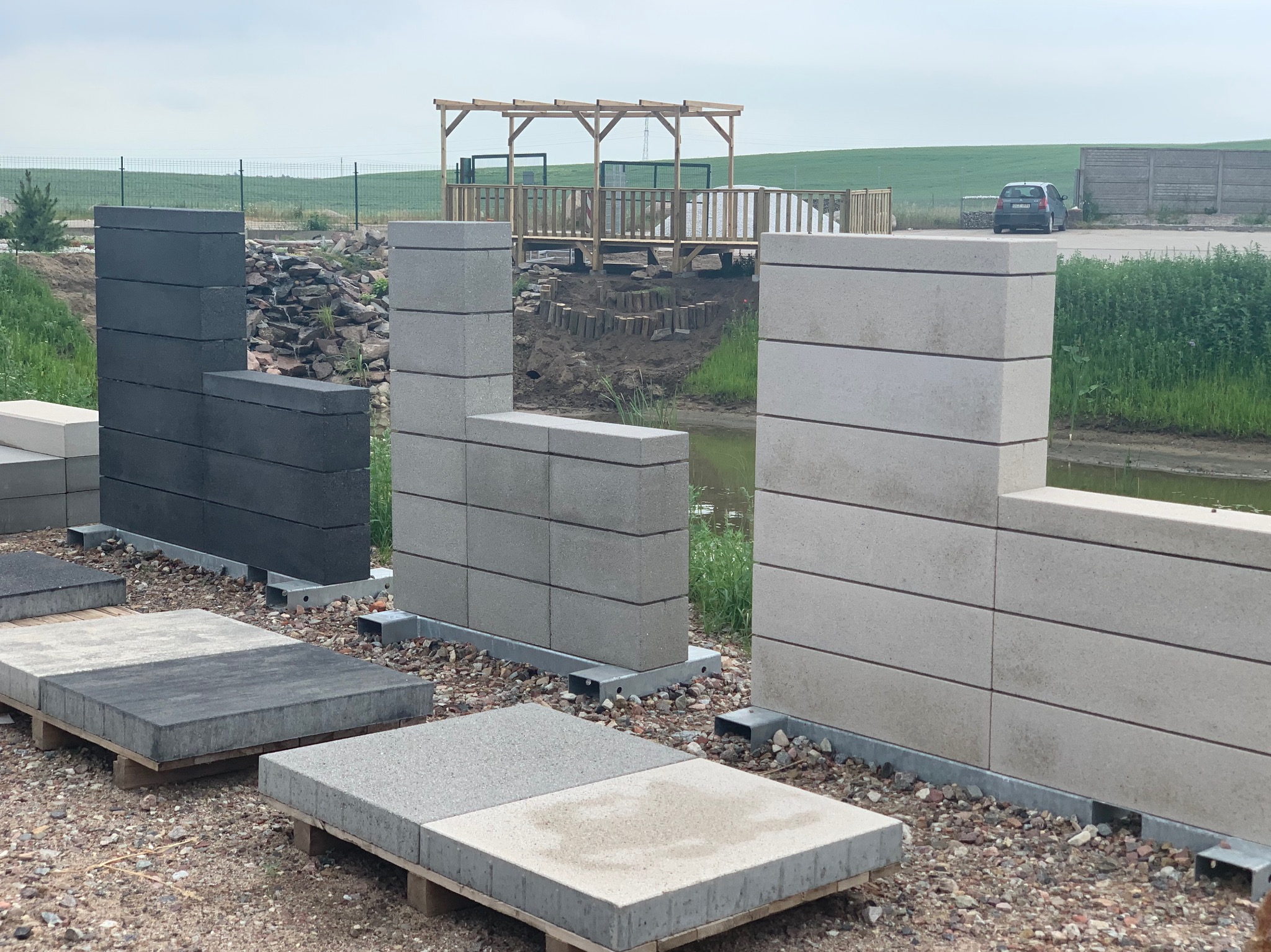 Ekspozycja elementów betonowych w różnych odcieniach szarości, ustawionych w formie niskich ścianek i płyt na paletach, z widoczną w tle drewnianą altaną i zielonym krajobrazem.