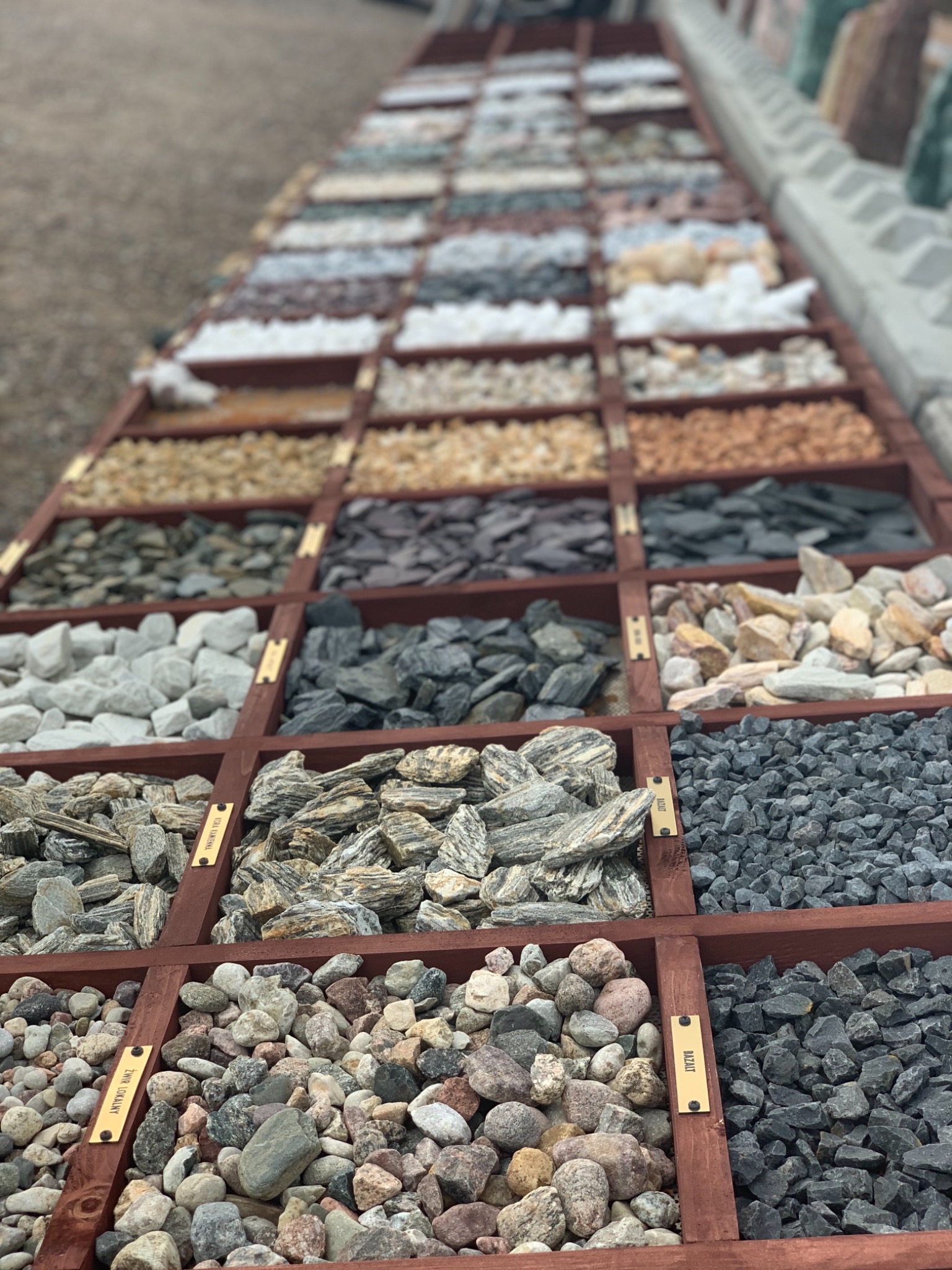 Ekspozycja różnorodnych kamieni dekoracyjnych i kruszyw w drewnianych przegródkach, z etykietami opisującymi rodzaje, prezentowana z perspektywy z góry, ukazująca bogactwo kolorów i faktur.