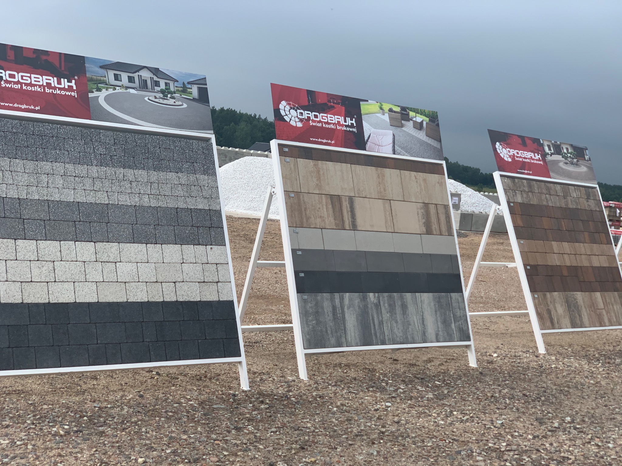 Ekspozycja trzech paneli z różnymi wzorami i kolorami kostki brukowej firmy Drogbruk na żwirowym podłożu, z widocznymi plakatami reklamowymi z logo i zdjęciami aranżacji.