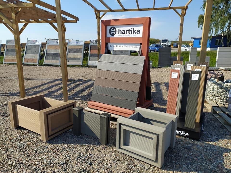 Ekspozycja zewnętrzna z donicami i panelami kompozytowymi w różnych odcieniach brązu i szarości, prezentowana na żwirowym podłożu z widocznym logo producenta.