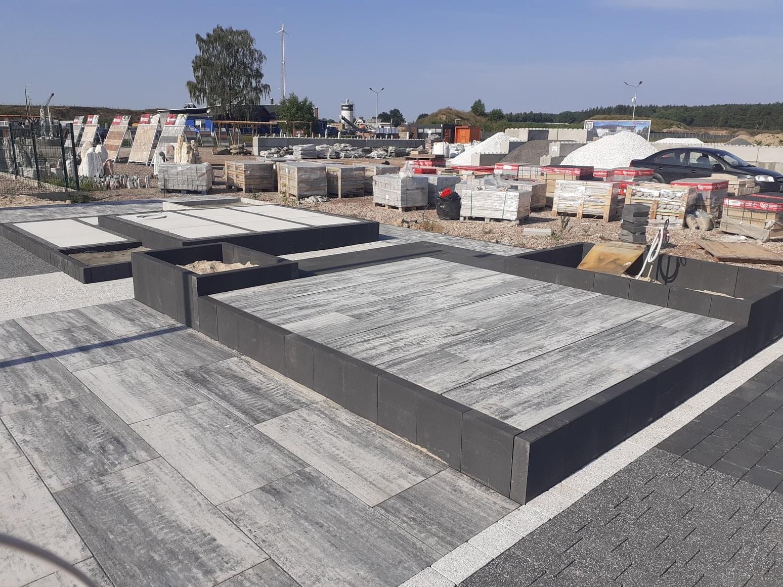 Ekspozycja różnych wzorów nawierzchni na placu budowlanym, z paletami materiałów w tle i wiatrakiem w oddali.