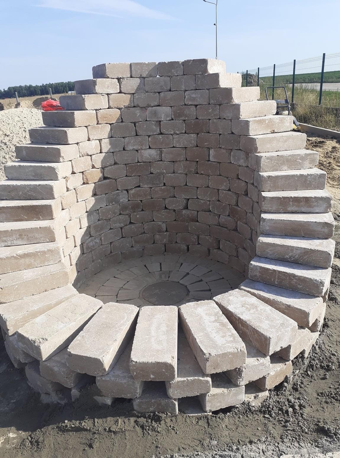 Okrągła konstrukcja z jasnych elementów nawierzchniowych, przypominająca studnię lub palenisko ogrodowe, w trakcie budowy, z widocznym tłem w postaci zieleni i nieba.