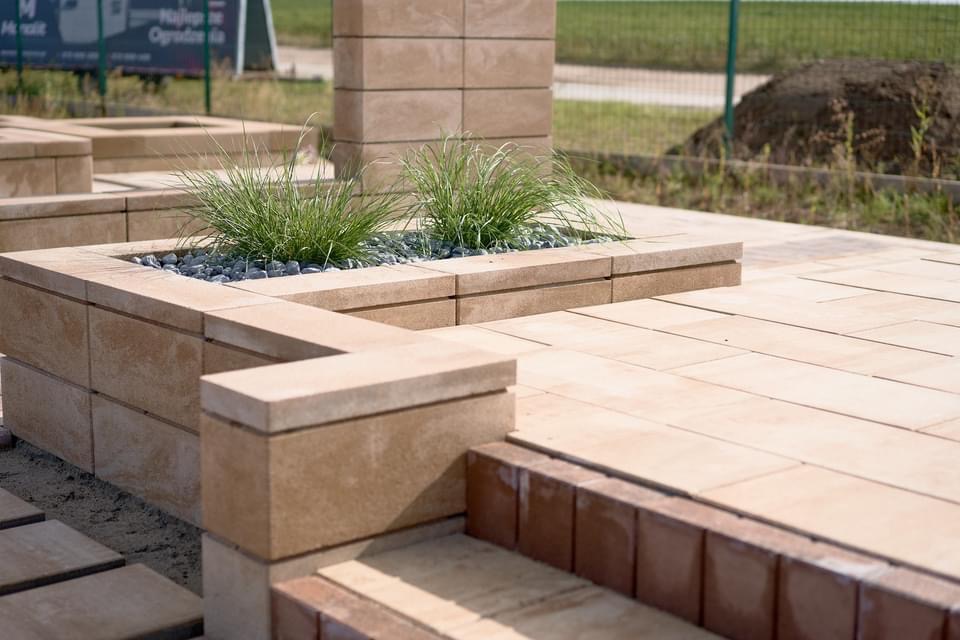 Wykończony taras z jasnej kostki betonowej z podwyższoną rabatą obsadzoną trawą ozdobną i wysypaną szarym żwirem, widoczne schody z czerwonej kostki.