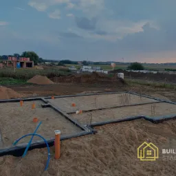 Przygotowany teren pod fundamenty domu jednorodzinnego z widoczną izolacją pionową, wystającymi rurami instalacyjnymi i częściowo zbrojeniem; w tle zabudowa jednorodzinna w trakcie budowy.