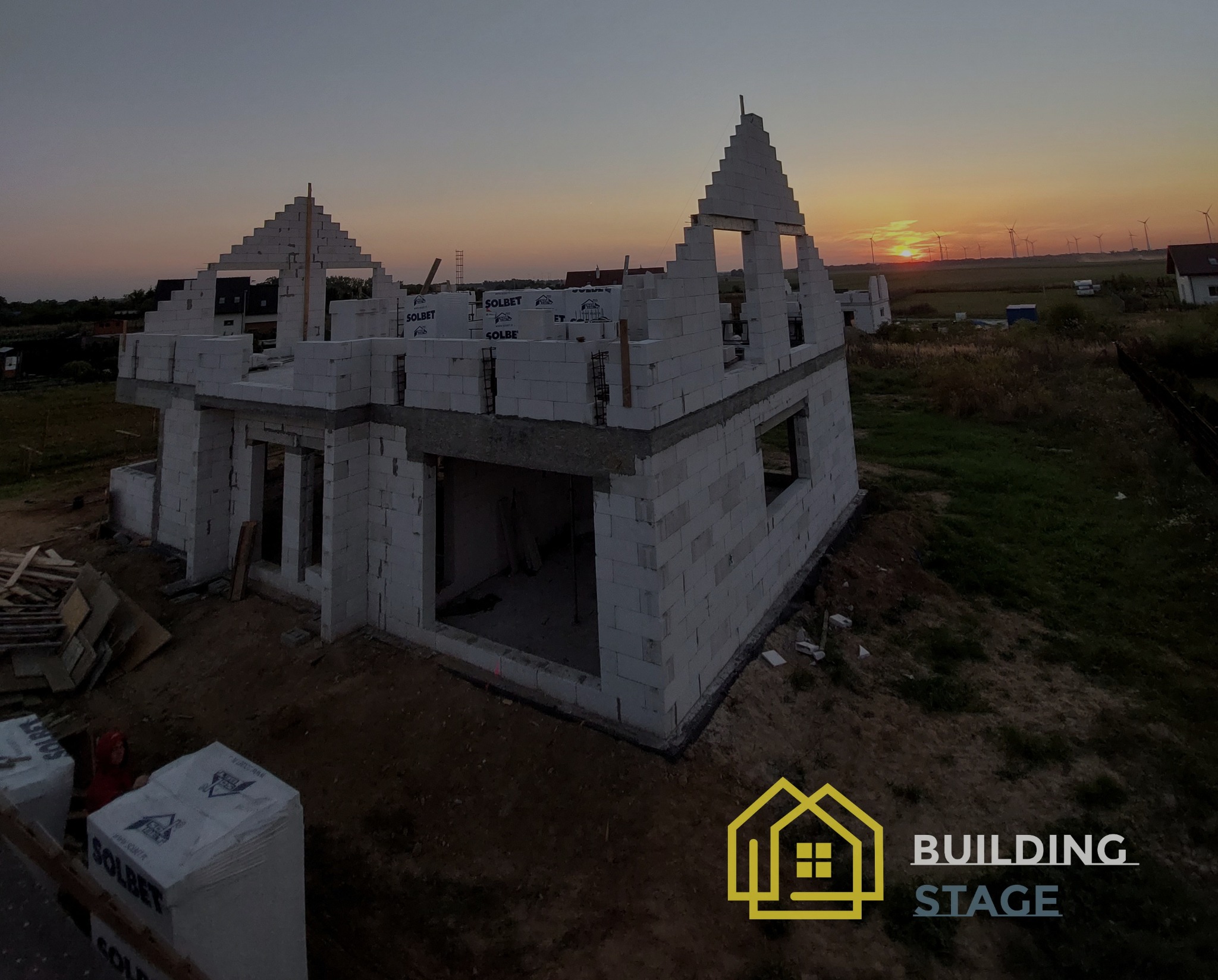 Wznoszenie ścian domu z betonu komórkowego Solbet na tle zachodzącego słońca i turbin wiatrowych.