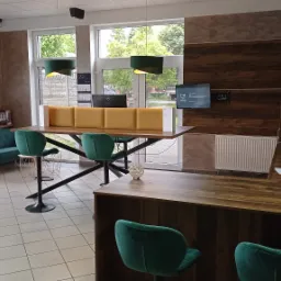 Wnętrze biura z recepcją: długa lada z blatem drewnianym, krzesła barowe w kolorze butelkowej zieleni, sofa w stylu retro, lampy wiszące i dekoracyjne ściany w odcieniach brązu i beżu.