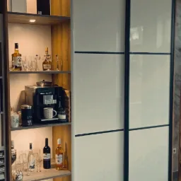 Wysoka szafa z przesuwnymi, białymi drzwiami z czarnymi ramami, po lewej stronie regał z drewnianymi półkami podświetlonymi punktowo, na których stoją butelki alkoholu, szklanki, ekspres do kawy...