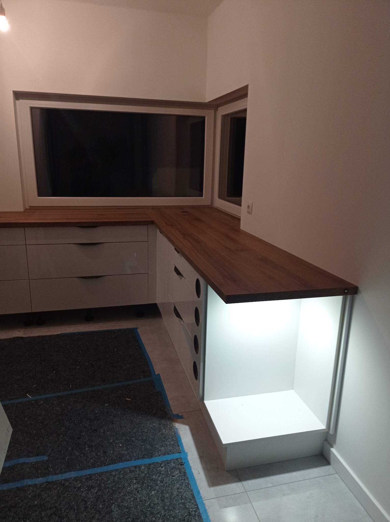 Nowoczesna zabudowa kuchenna z drewnianym blatem pod oknem, białe fronty szafek z ciemnymi uchwytami, oświetlenie LED w otwartej szafce, widoczna podłoga zabezpieczona taśmą malarską.