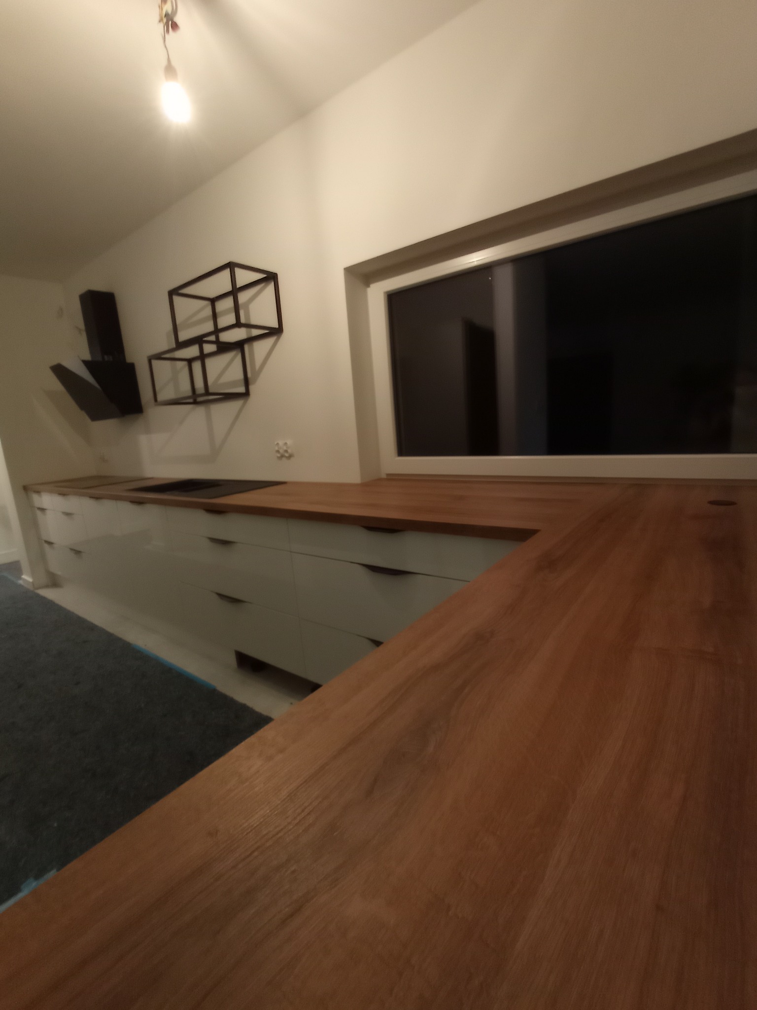 Nowoczesna kuchnia z białymi, lakierowanymi frontami i blatem z ciepłego drewna, oświetlona pojedynczą żarówką, z czarnym okapem i geometryczną dekoracją ścienną.