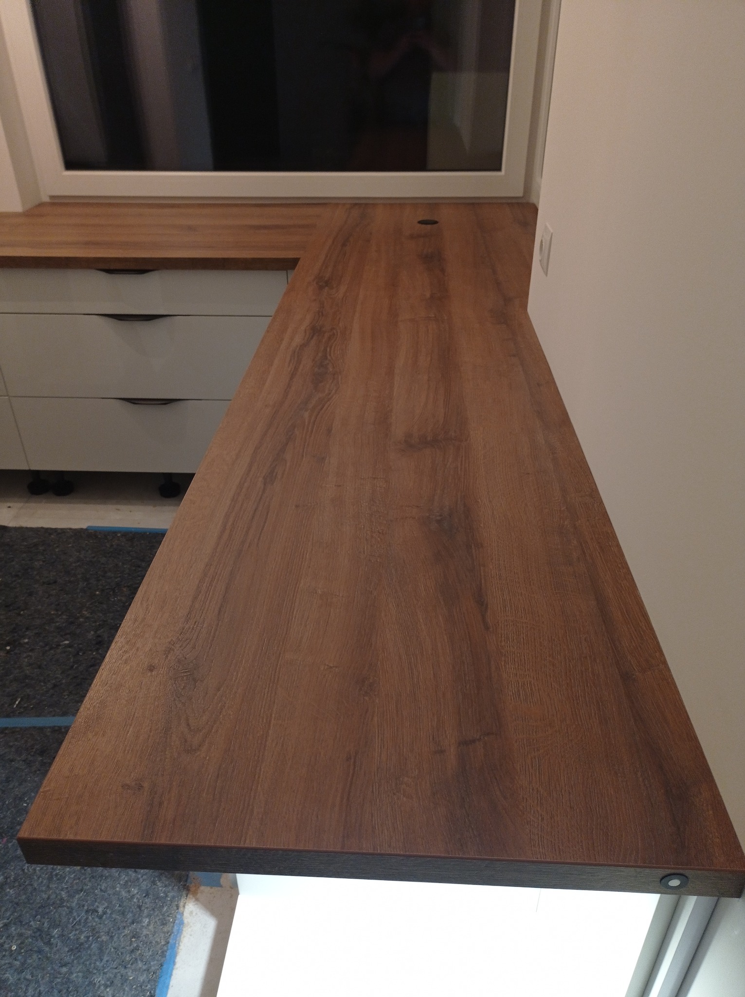 Brązowy blat drewniany z otworem, zamontowany przy oknie nad białymi szafkami z szufladami, widoczny fragment szarej wykładziny.