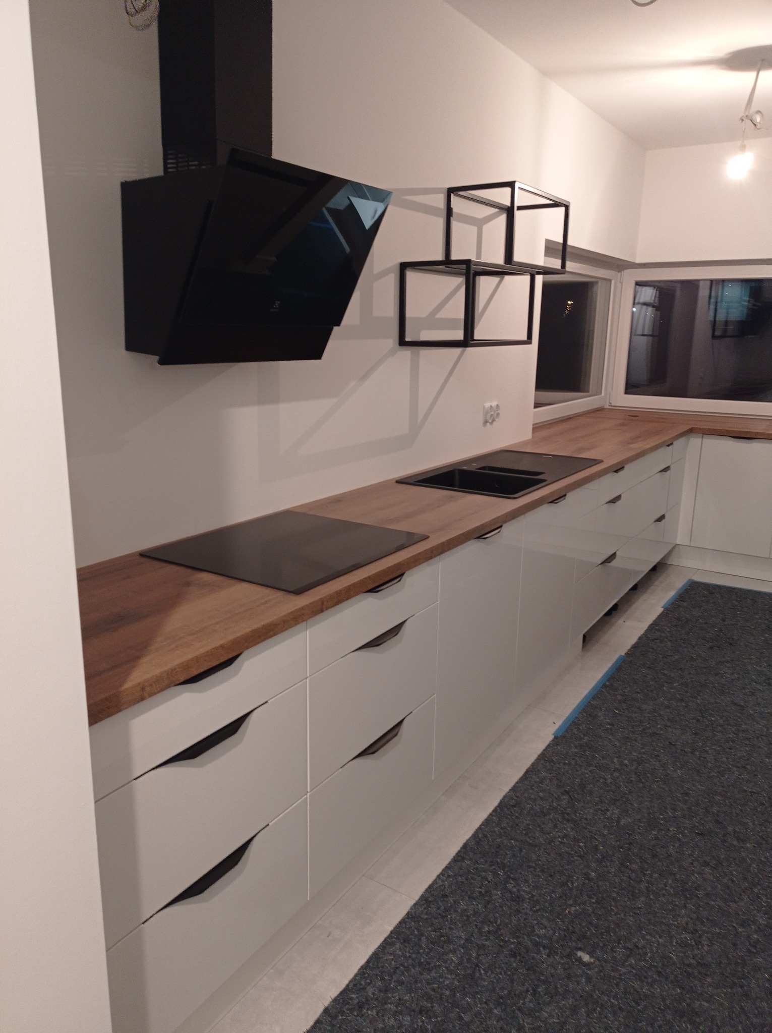 Nowoczesna kuchnia z białymi szafkami z frezowanymi uchwytami, drewnianym blatem, czarną płytą indukcyjną i okapem oraz dekoracyjnymi półkami w kształcie kostek.