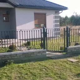 Czarna, metalowa balustrada z motywem greckim, osadzona na murku z cegły, otaczająca posesję z domem w tle. Brama wejściowa z tego samego wzoru.