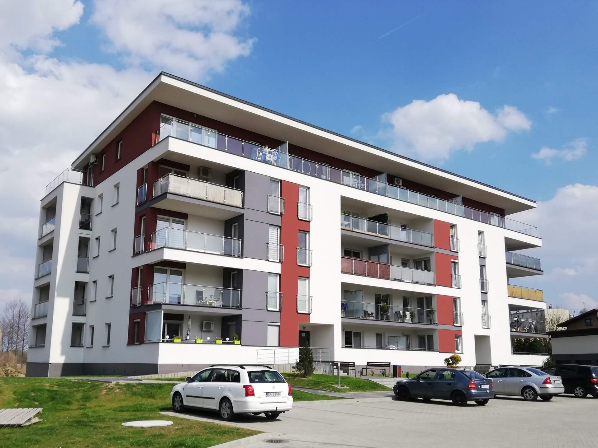 Nowoczesny, wielopiętrowy budynek mieszkalny z balkonami wykończonymi szkłem, pomalowany na biało, czerwono i szaro, z zaparkowanymi samochodami na pierwszym planie i błękitnym niebem w tle.