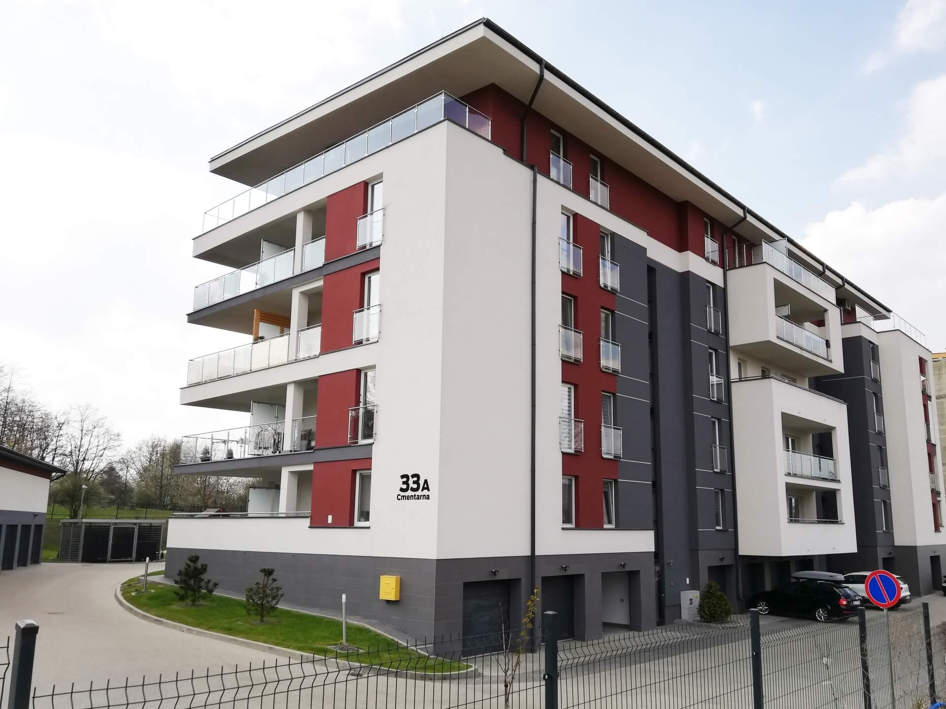 Nowoczesny, wielopiętrowy budynek mieszkalny z balkonami wykończonymi szkłem, pomalowany w kolorach białym, szarym i ceglastym, z widocznym numerem budynku '33A' i nazwą ulicy 'Cmentarna'.