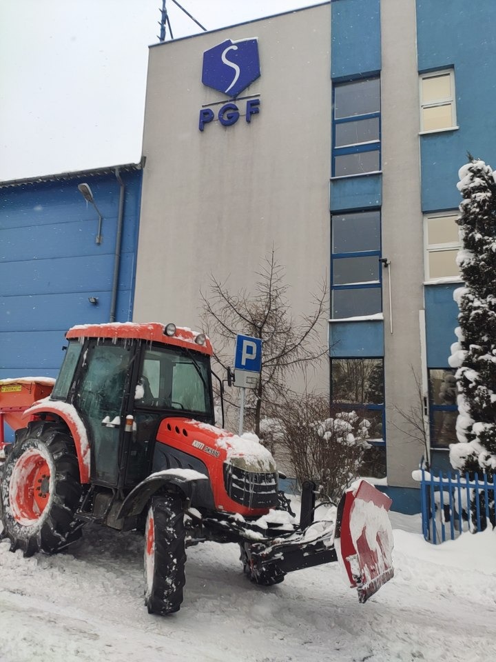 Pomarańczowy traktor z pługiem śnieżnym, częściowo pokryty śniegiem, stoi na ośnieżonym parkingu przed budynkiem z logo PGF i znakiem parkowania.