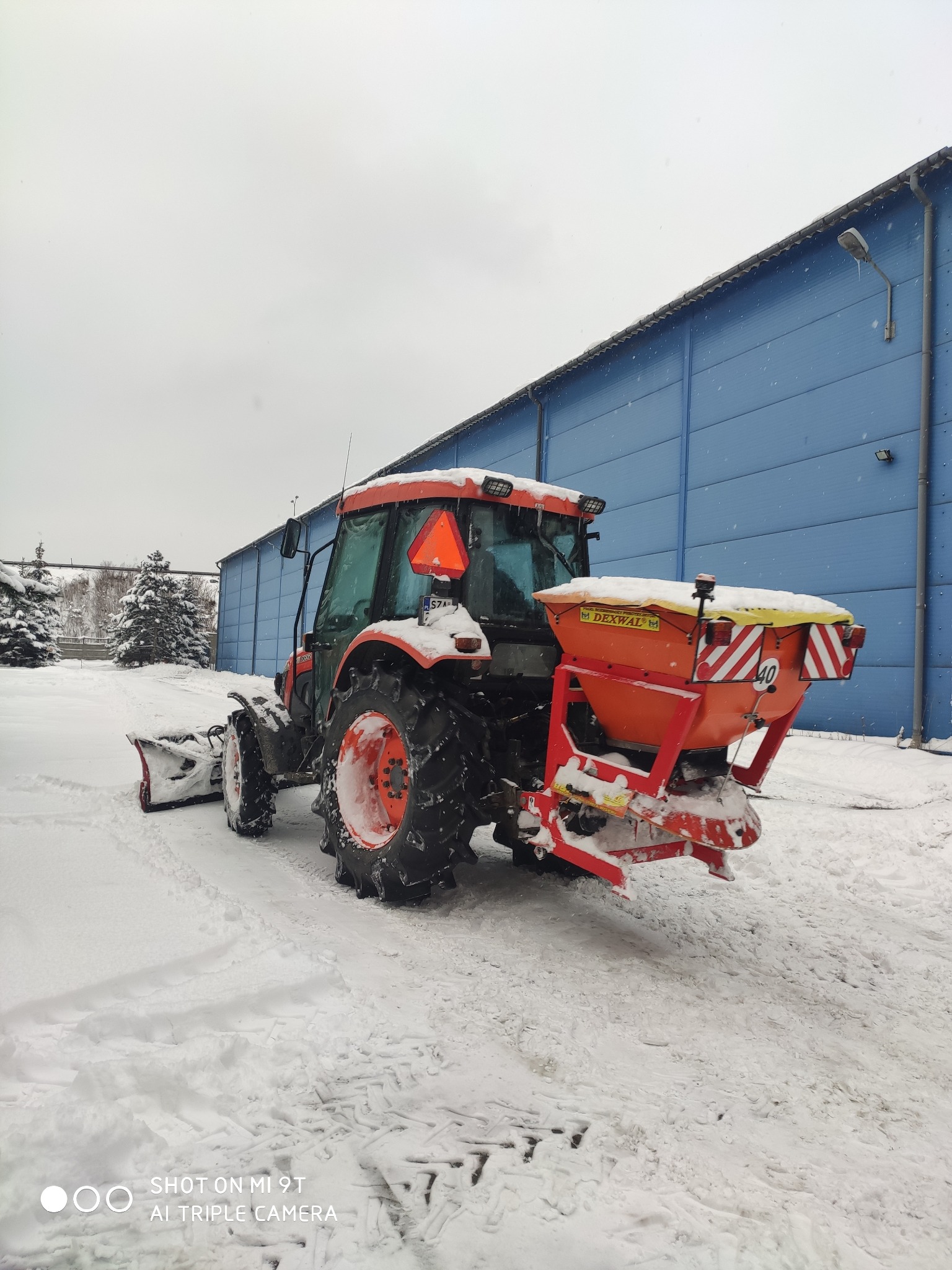 Pomarańczowy traktor z pługiem i rozsiewaczem soli/piasku, stojący na ośnieżonym placu przed niebieskim budynkiem przemysłowym w pochmurny dzień.